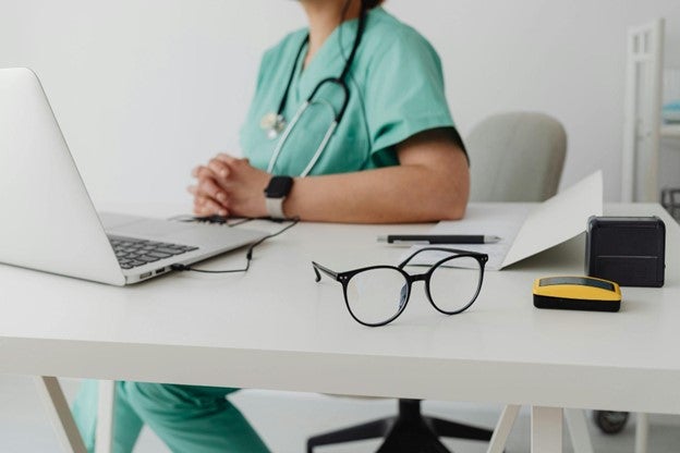 Healthcare professional in scrubs using a laptop at a desk with glasses and stationery.