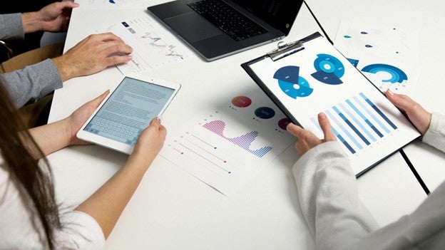Three individuals analyzing data and graphs on digital tablets and printed sheets at a white table.
