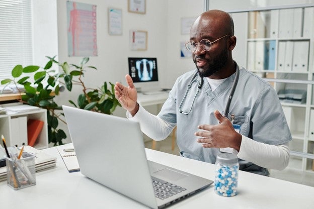 Doctor in scrubs with stethoscope gesturing during a video call on a laptop in an office.