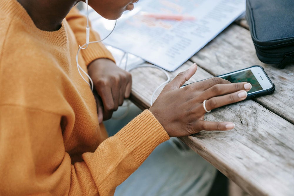 Person in a sweater using a smartphone with earphones, resting on a wooden table.